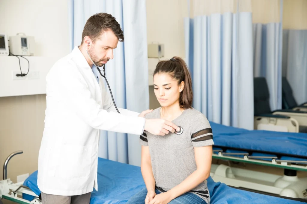 Male doctor listening to young female patient’s heart with stethoscope in hospital room for immigration medical exam Houston.