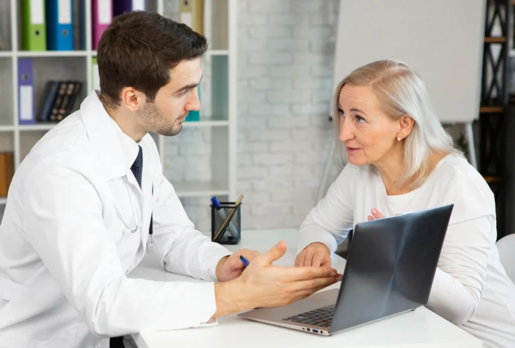 Dedicated male doctor explaining to senior female patient using laptop in modern office during USCIS immigration medical physical consultation.
