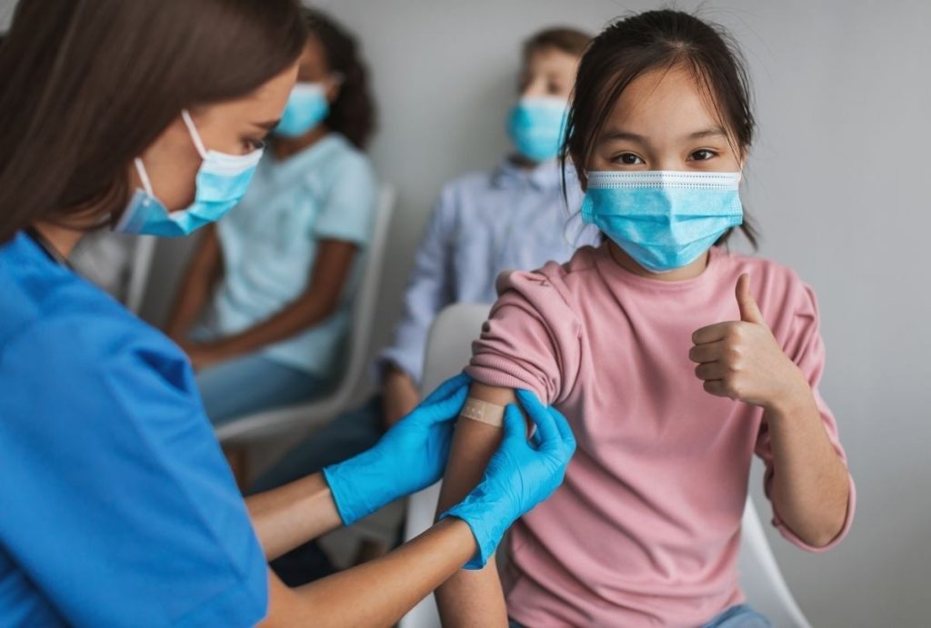 Healthcare worker applying bandage after vaccine as child gives thumbs-up during USCIS immigration medical physical exam.