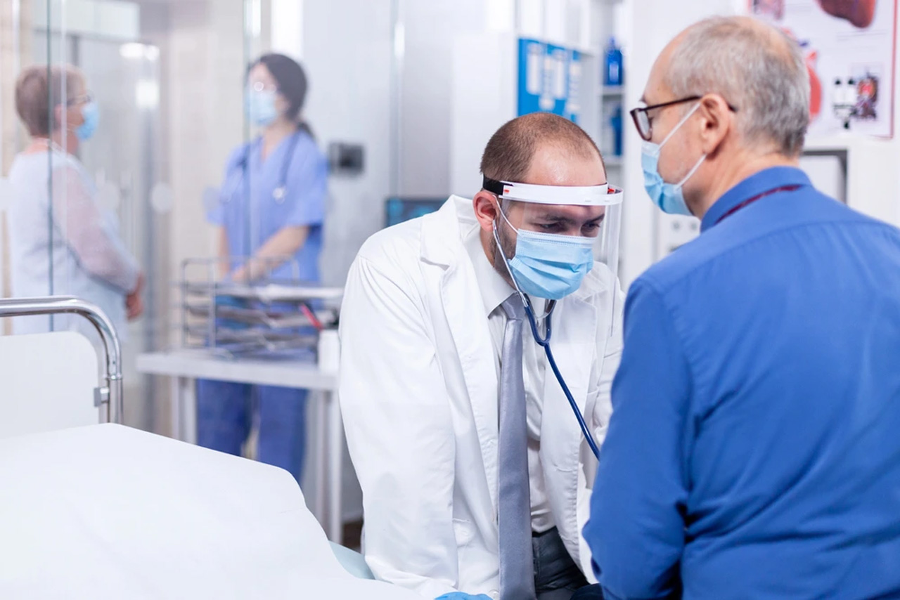 USCIS immigration medical exam where doctor in protective gear listens to senior patient’s chest while clinical team supports evaluation