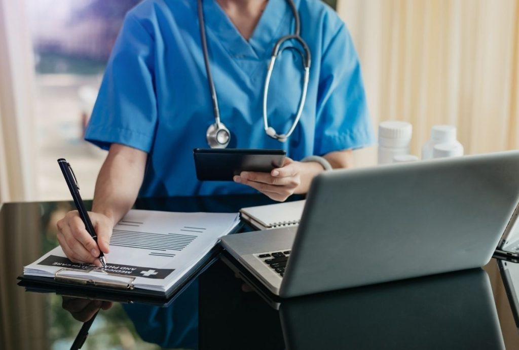 Doctor in scrubs reviewing records and writing notes during low cost immigration medical exam Houston with digital tools.