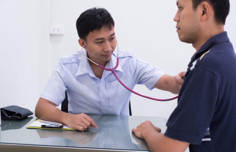 Male doctor conducting an immigration medical physical Houston exam using a stethoscope to check a patient's heart.