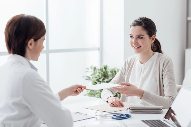 Doctor in white coat reviews paperwork with smiling patient after her required USCIS immigration medical exam in a bright office.