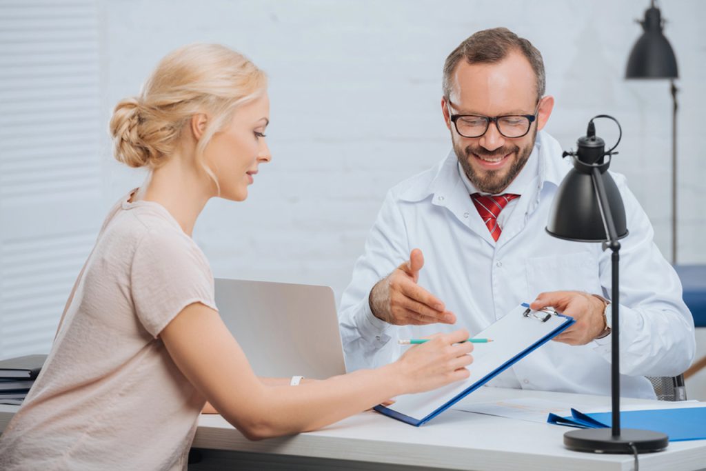A female patient signs forms presented by a smiling doctor during a USCIS immigration medical physical consultation.