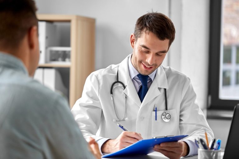 A friendly immigration doctor Houston smiles while completing a patient's medical history form on a blue clipboard.