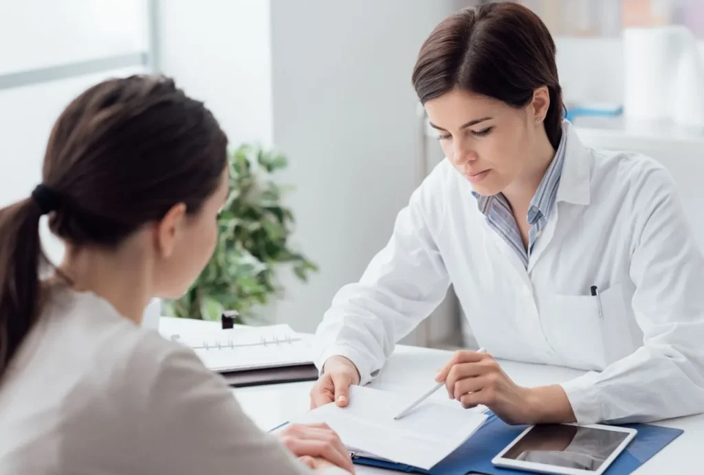 Woman doctor with a white coat points to a form during a patient's essential green card medical exam consultation.