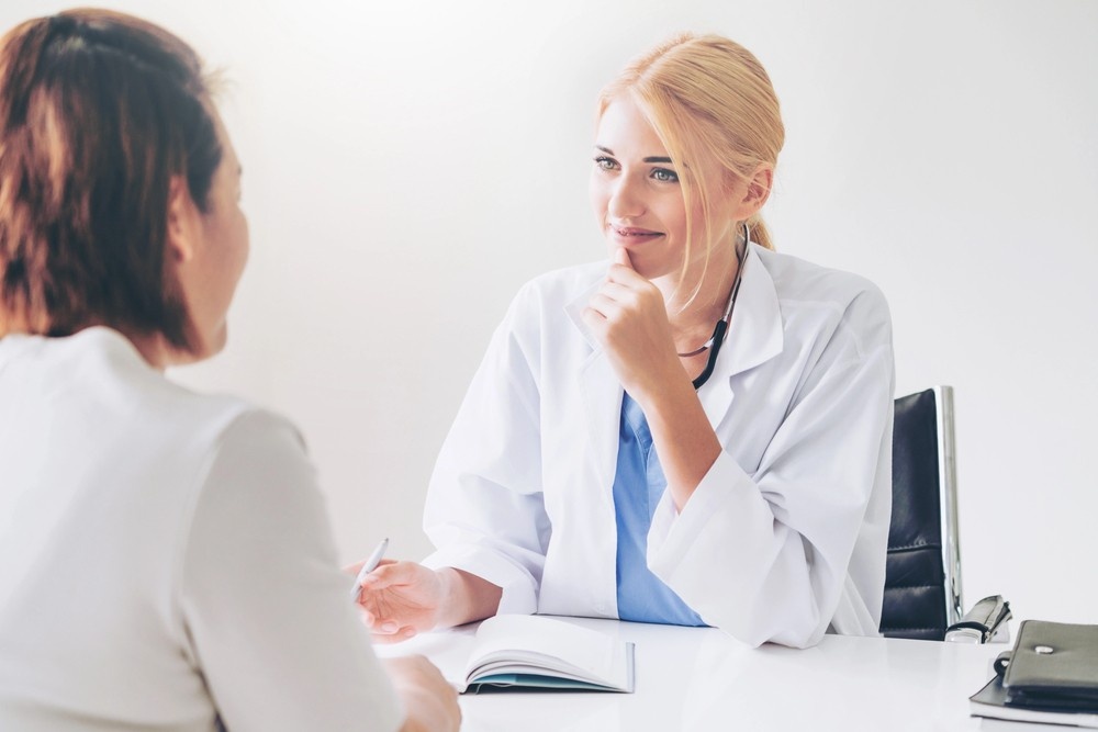 A blonde immigration doctor Houston specialist listens intently to a patient across a white desk during a checkup.