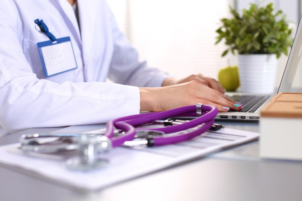 A doctor typing on a laptop near a purple stethoscope while processing the cheapest immigration medical exam Houston.